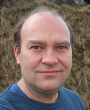 A white man wearing a blue T shirt looks directly at the camera, smiling slightly.